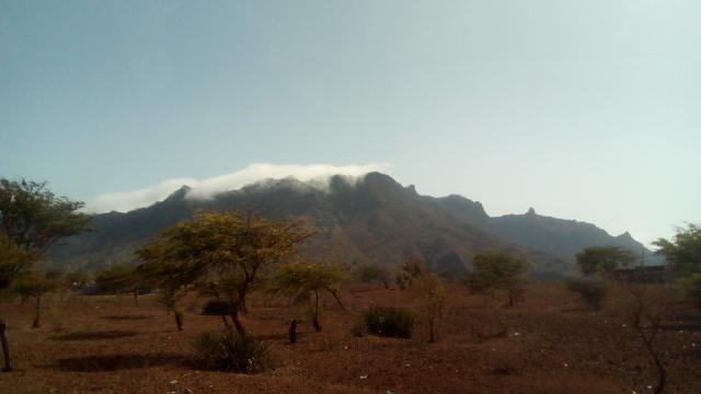 Montanha com nuvens ao fundo de uma paisagem de terra árida com algumas árvores